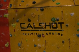 Yellow indoor climbing wall with multiple coloured climbings holds and the centres logo "Calshot Activity Centre" written in black across the middle of the wall.