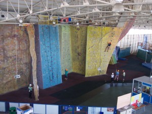 Pan view of Calshot climbing walls.  Shows several of the centres climbing walls, each of which is in a different colour, with multiple coloured climbing holds.  The yellow at the far right of the image has a person climbing half way up to the top.