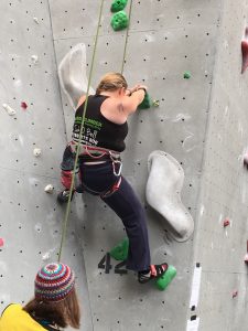 View of me on a grey wall with green holds, using my left foot to balance on the wall beside me