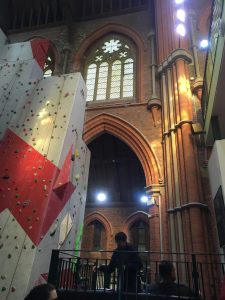 interior image of Manchester Climbing Centre, looking up to a large stain glassed window, above a brick archway, exposed brick and pillars are seen along with a light coloured climbing wall to the left, with different coloured holds and decorative features.