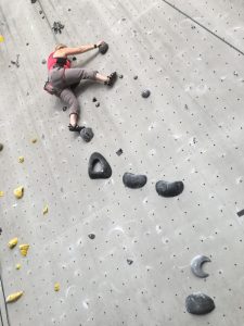 Image of T climbing on a grey wall with black holds, while being positioned in a capital K position.