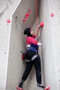 Anoushé is climbing within a ‘chimney’ at Ratho Climbing centre, she is surrounded by pink jug and bicycles handlebar type holds, her left hand is about head hieght on a hold and she is just moving her right arm, which is covered in white tape at the end onto a similar hold