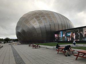 Photograph shows a round ball like building on the left which is Glasgow IMAX with a large screen on the right of the photograph, with a small green area in front of it