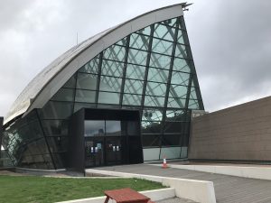 Photograph of the entrance to The Glasgow Science Museum, shoring what looks like the top half of a letter C shaped building, the glass front of the building sat to the right of the large screen