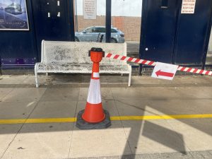 Photo of the inside of Fareham Bus Station, a large metal timetable board is on the left and infront is a orange cone with red and white tape and a sign pointing left to state that this is the direction to walk.  In the background past the tape is a metal bench.