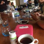 Image of Members of the group sat around a large table with phones, iPads and other tech sat on the table with numerous cups of tea, coffee and water with Molly stood at the head of the table showing her iPad:
