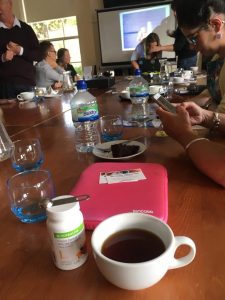 Image of Members of the group sat around a large table with phones, iPads and other tech sat on the table with numerous cups of tea, coffee and water with Molly stood at the head of the table showing her iPad: