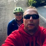 Photograph shows a ‘selfie’ of Simon smiling at the camera, with me, Tee sat behind him on the left. The photo is of us both sat on the Tandem, but the bike is not in the shot.