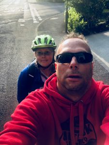Photograph shows a ‘selfie’ of Simon smiling at the camera, with me, Tee sat behind him on the left. The photo is of us both sat on the Tandem, but the bike is not in the shot.