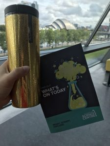 Photograph of my gold Starbucks travel mug with the guide book to The Glasgow Science Centre with the SEC Armadillo building behind across the river Clyde.
