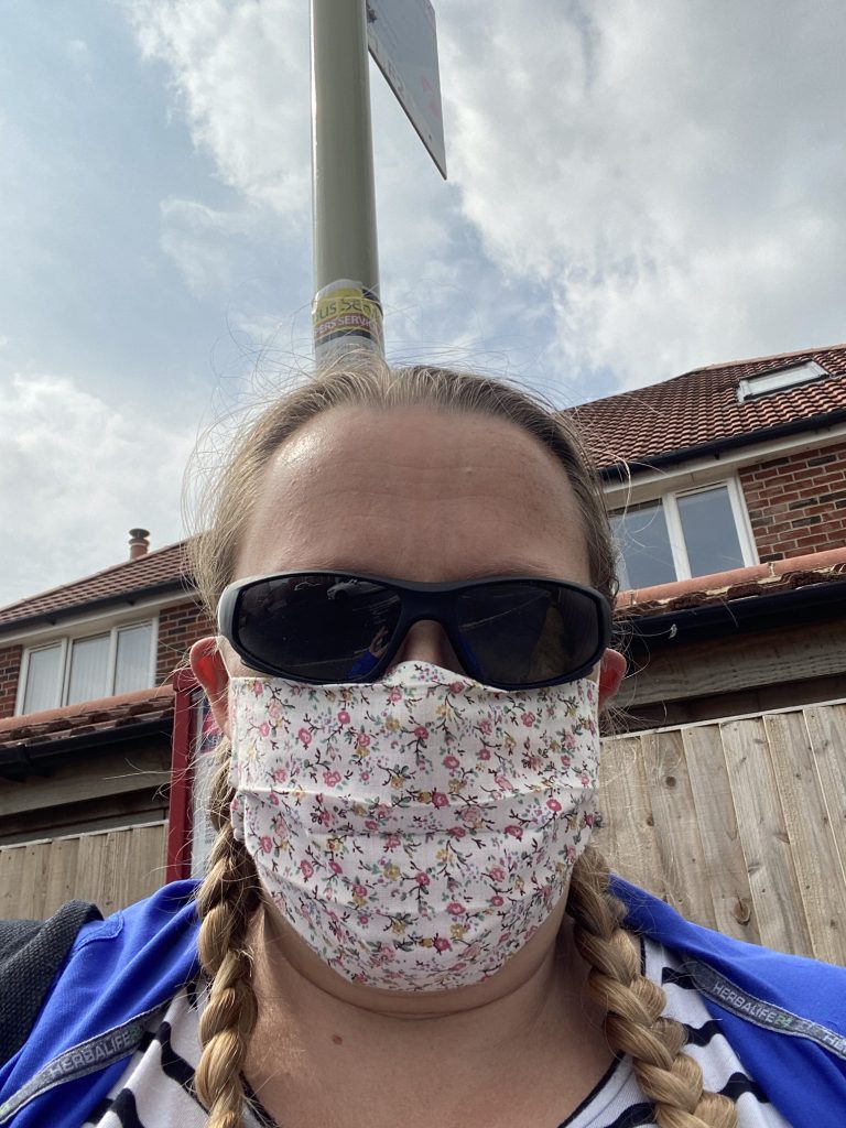 Selfie photo of me wearing a floral face mask and dark sunglasses with the pole for the bus stop behind me