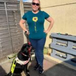 Photograph of Tee on the patio with a cream garden wall behind, with Guide dog fizz sat on the left in her harness . Both looking at the camera.