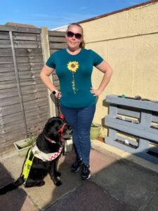 Photograph of Tee on the patio with a cream garden wall behind, with Guide dog fizz sat on the left in her harness . Both looking at the camera.